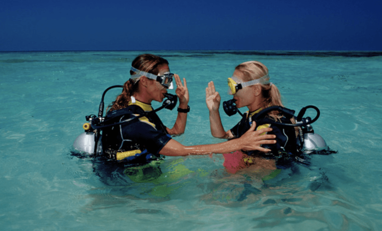 Two scuba divers wearing wetsuits and diving gear stand in clear, shallow turquoise water, facing each other and making hand signals, possibly communicating or practising dive skills.