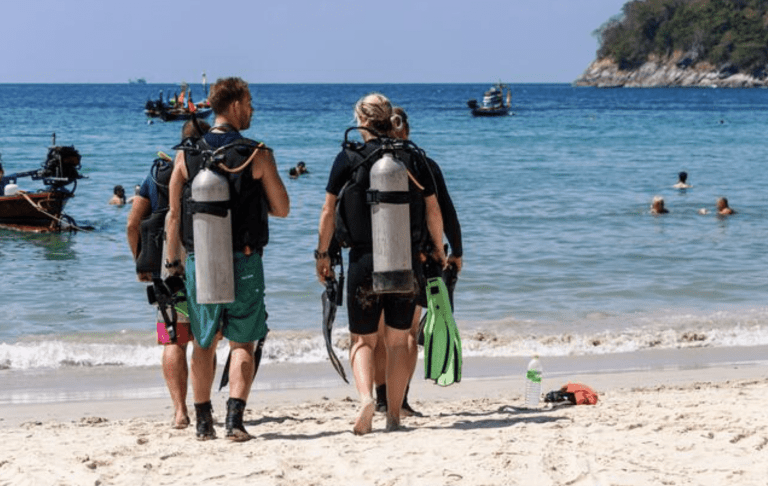 Three people in scuba gear walk towards the sea on a sandy beach, carrying fins and snorkels. Other people are swimming and small boats are anchored offshore under a clear sky.