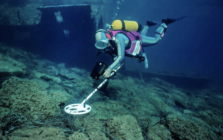 A scuba diver wearing a wetsuit and carrying an oxygen cylinder uses an underwater metal detector to search the sea floor near a sunken shipwreck.