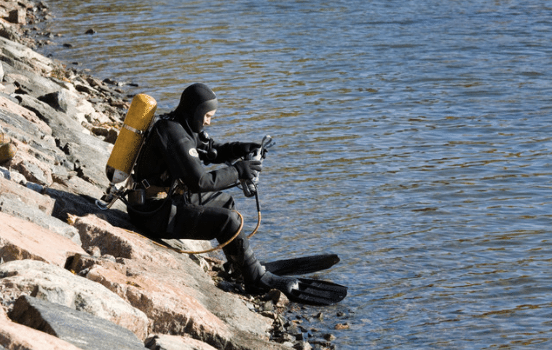 A scuba diver in a black wetsuit and yellow oxygen cylinder sits on rocky shore, adjusting equipment while facing a body of water.