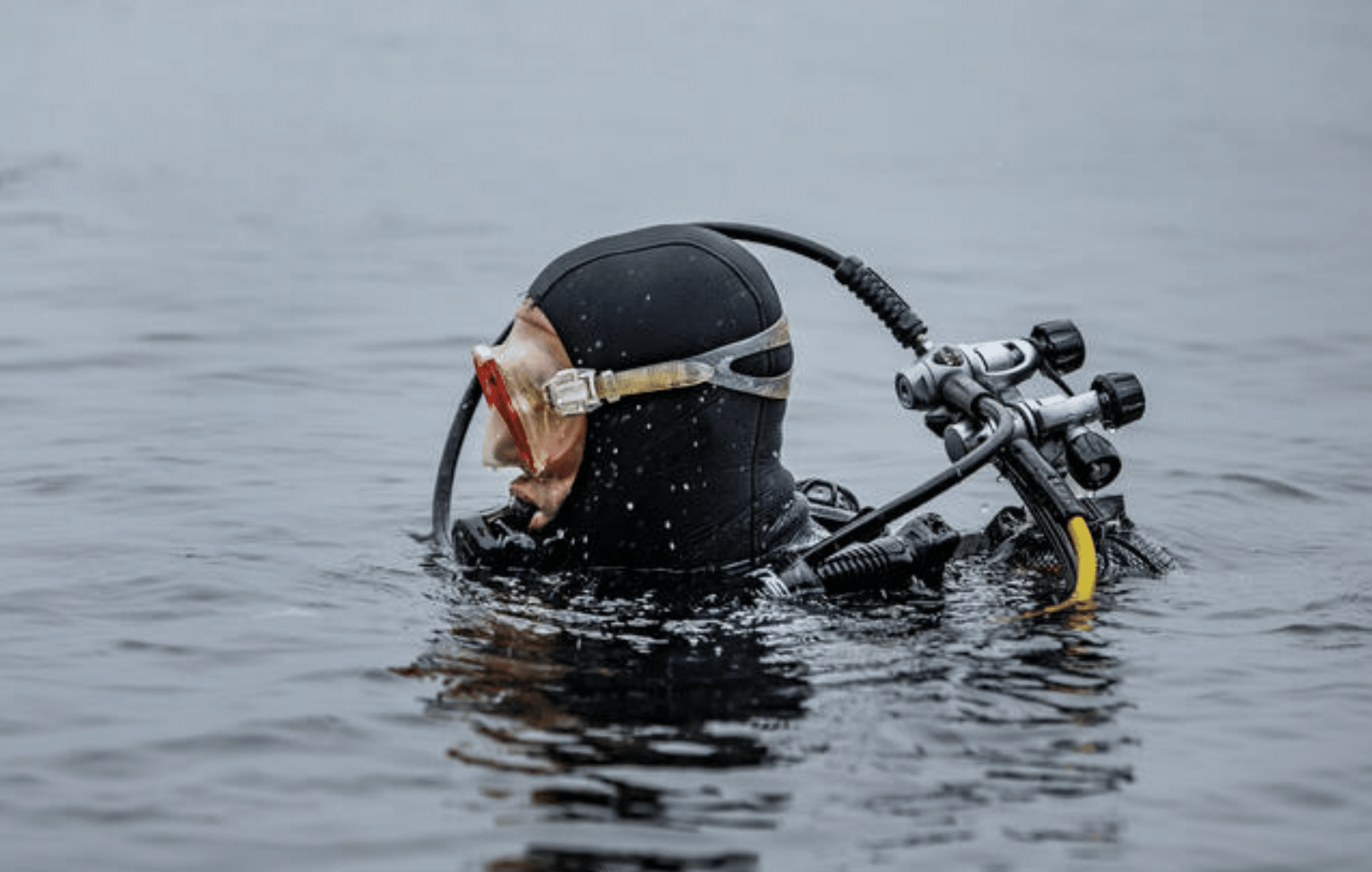 A scuba diver in a black wetsuit and red goggles is partially submerged in water, wearing a full set of diving kit with an oxygen cylinder on their back.